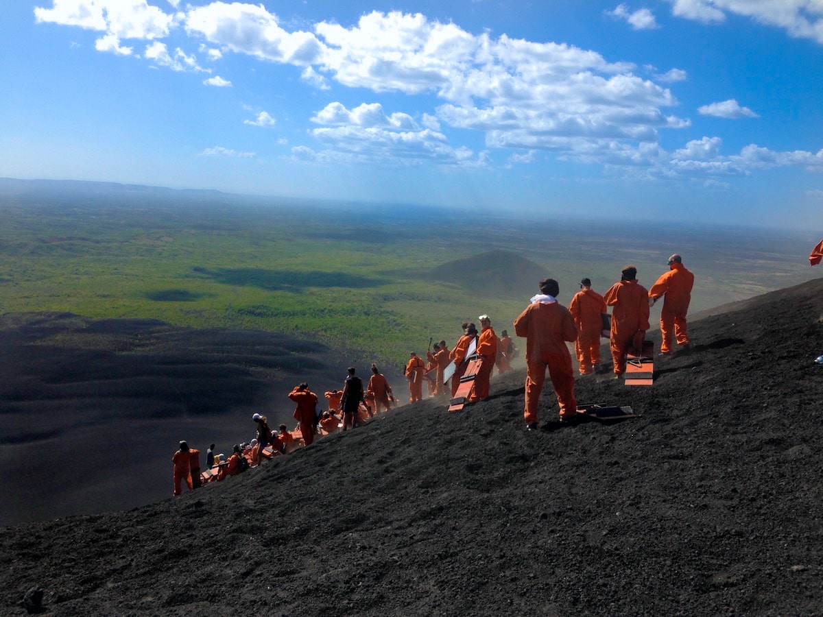 A tour group in orange jumpsuits hold their boards on a volcano. 