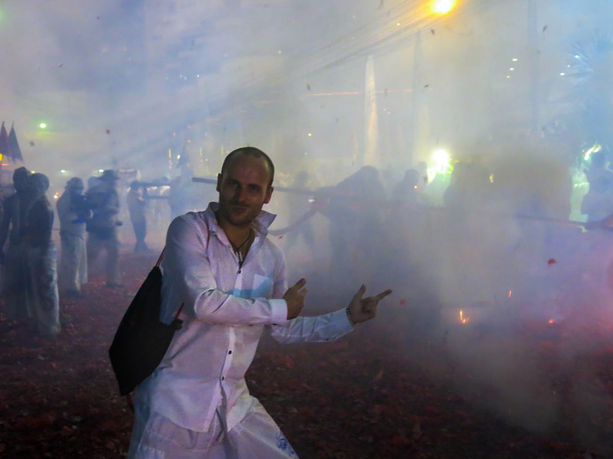Man in white is pointing at a street parade with fireworks and smoke. 