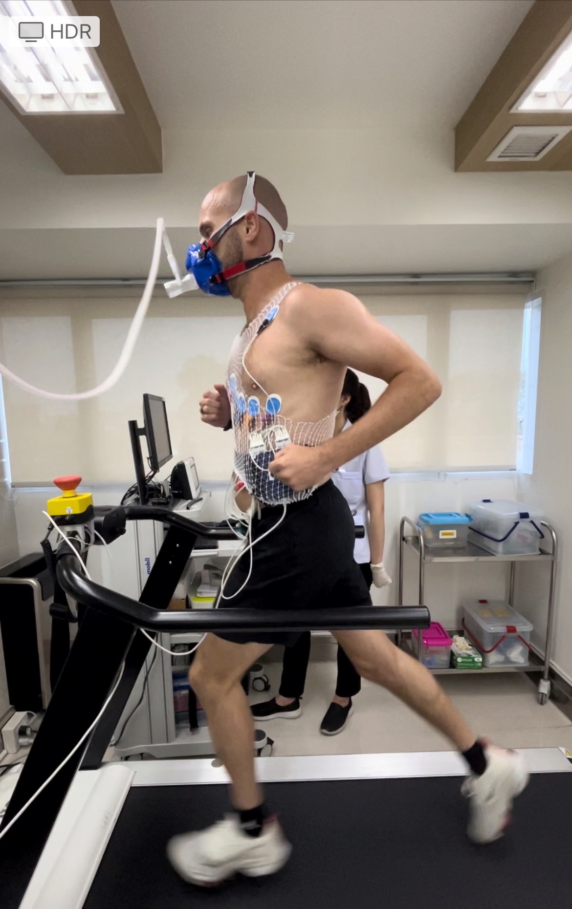 A shirtless man runs on a treadmill with a mask over his face and wires attached to his body from a machine