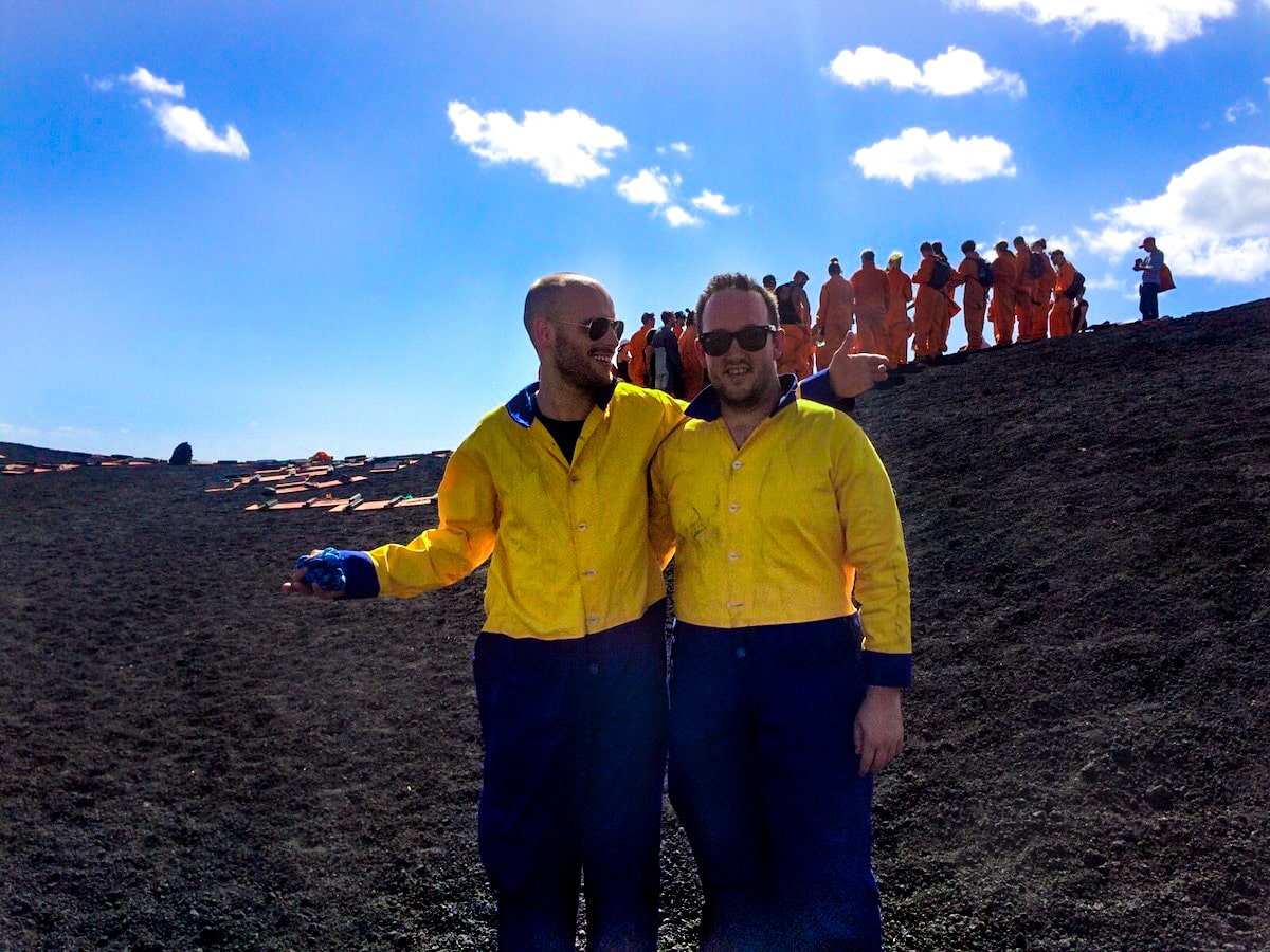 Two men in a yellow and blue jumpsuits stand on black volcanic ash in front of a tour group in orange jumpsuits. 