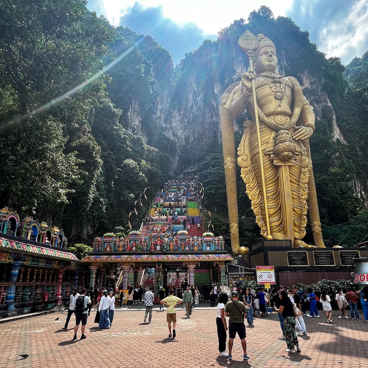 Visitors at the Batu Caves in Malaysia, with the towering golden statue of Lord Murugan and the vibrant, colorful staircase leading up to the caves set against a backdrop of lush green cliffs