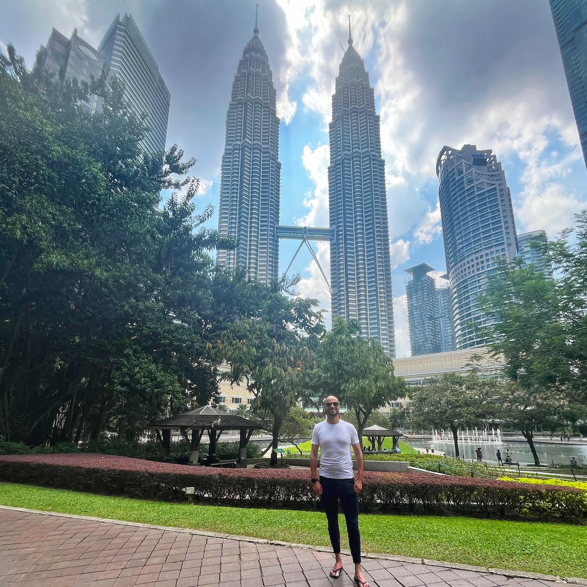 A man standing in a park with the Petronas Twin Towers in Kuala Lumpur, Malaysia, towering in the background under a partly cloudy sky. The lush greenery of the park contrasts with the modern skyscrapers.