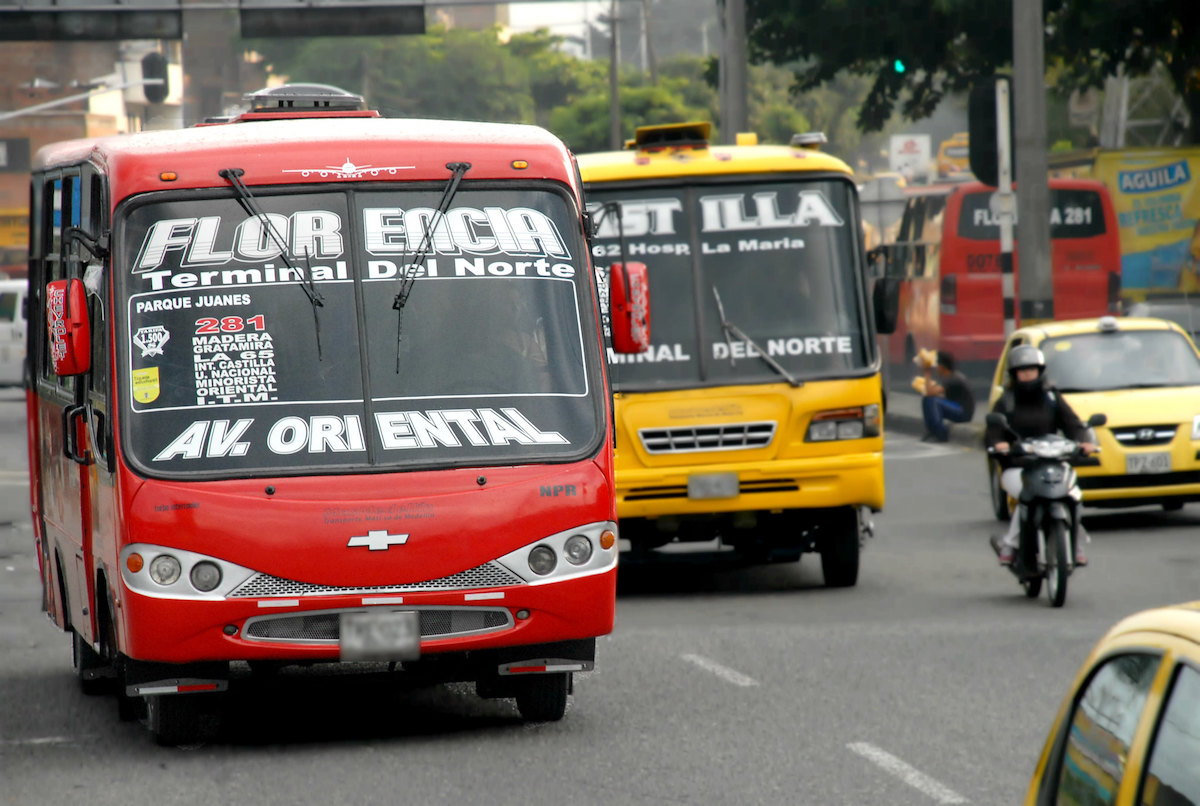A red bus and yellow bus drive alongside a motorbike in Medellin, Colombia. 