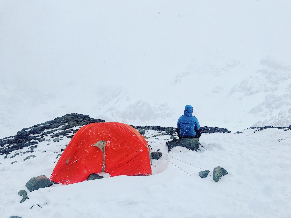 A solitary man in blue looks out into the wilderness in a snowy harsh climate, next to an orange tent
