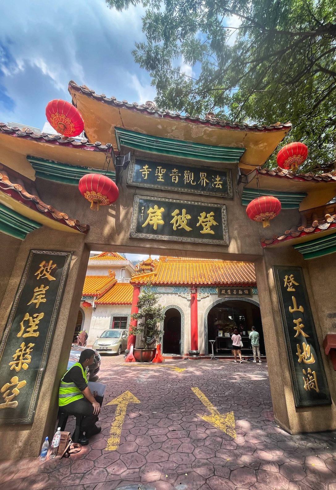 Entrance to the Dharma Realm Guan Yin Sagely Monastery in Kuala Lumpur, Malaysia, featuring traditional Chinese architectural elements, red lanterns, and gold inscriptions. Two people are sitting near the entrance, and visitors can be seen entering the monastery.