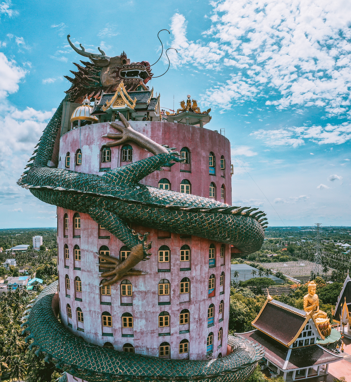 A large pink tower building with a dragon statue wrapped around it towers over a Buddha statue below. 