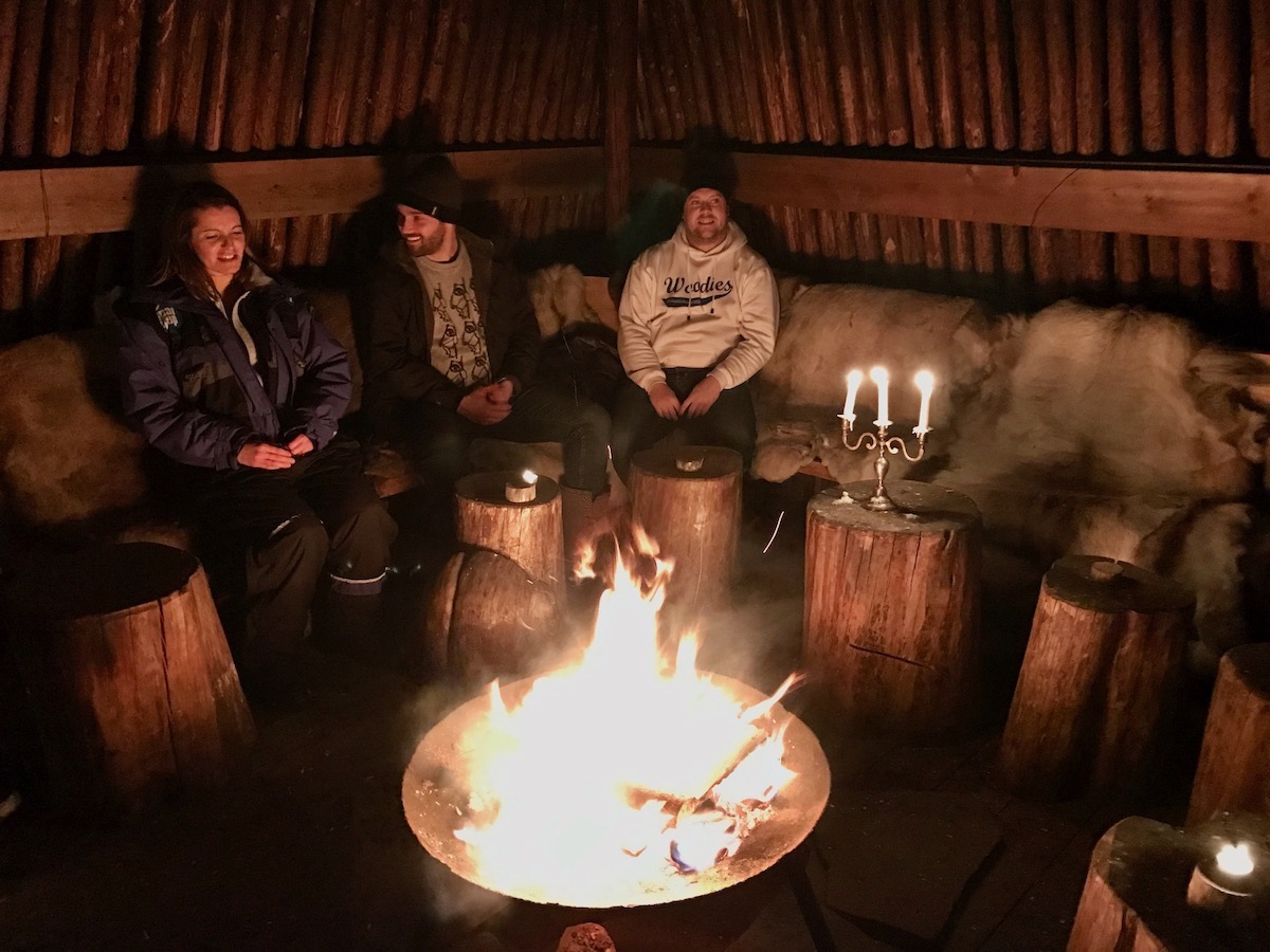 Two men and a woman sit around a log fire in a cabin in Kirkenes, Norway