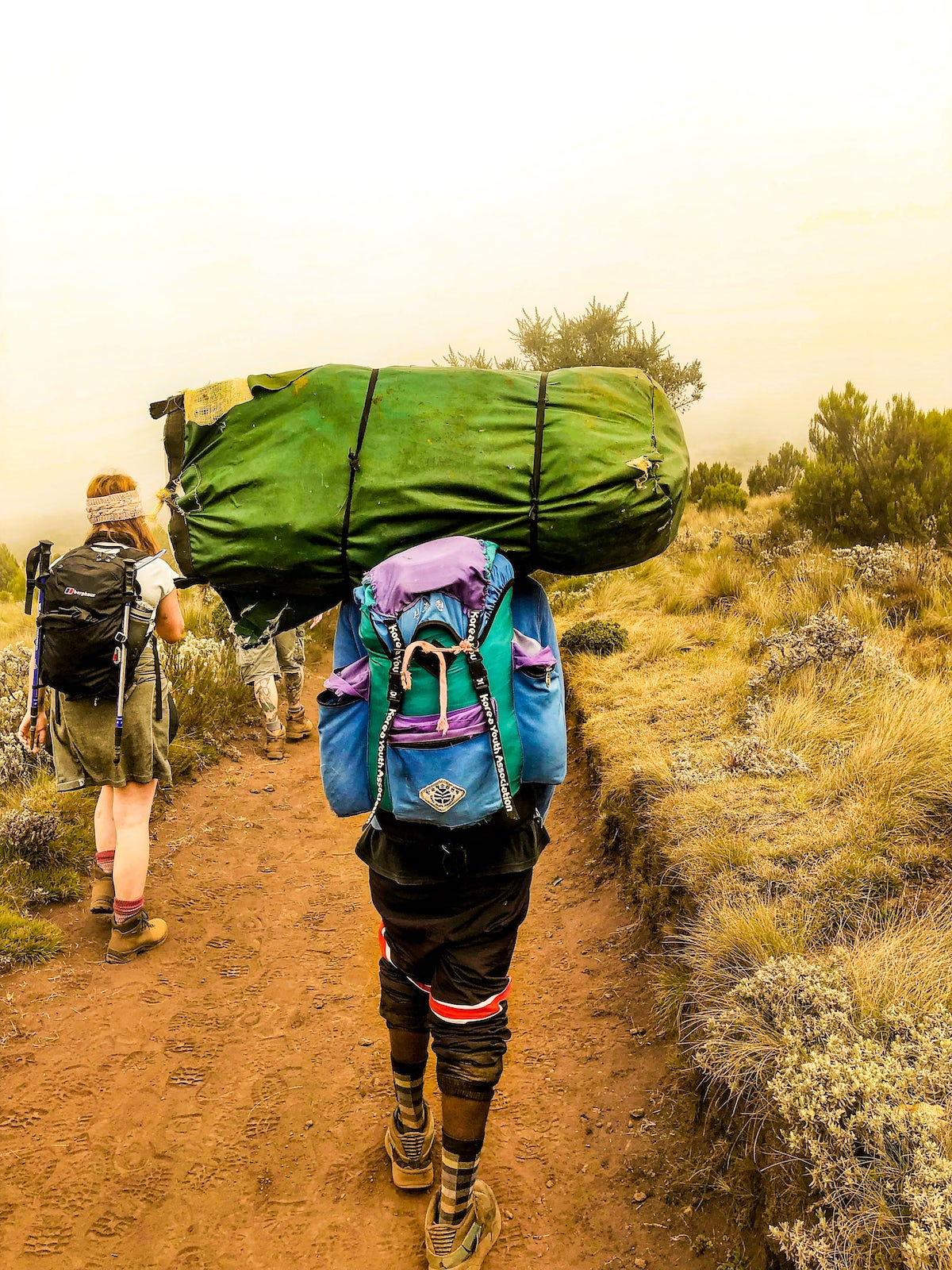 A porter on a mountain carries a large green bag on his head.