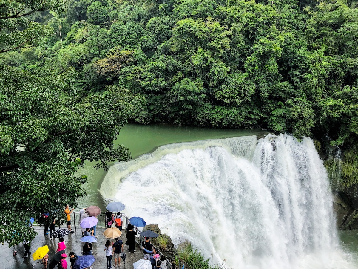 Tourists hold up their umbrellas near a large flowing waterfall under an emerald green lake and trees surrounding it