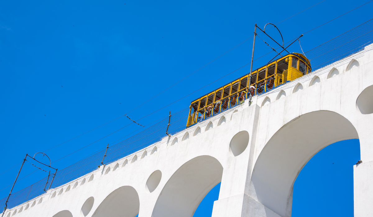 A yellow tram travels over a white arch on a bright blue sky day in Lapa, Rio de Janeiro, Brazil.