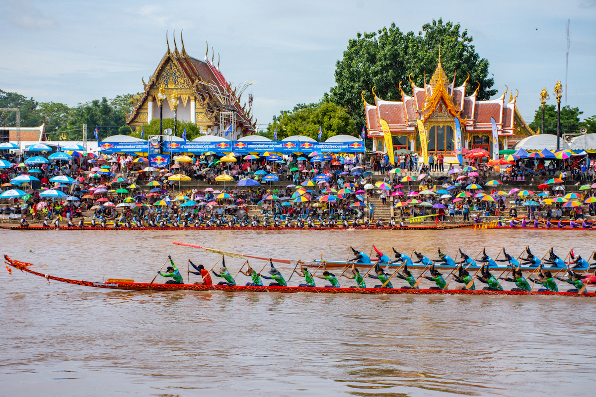 Two long boat teams race against each other in a famous Thailand festival with a packed crowd watching in front of two golden temples.