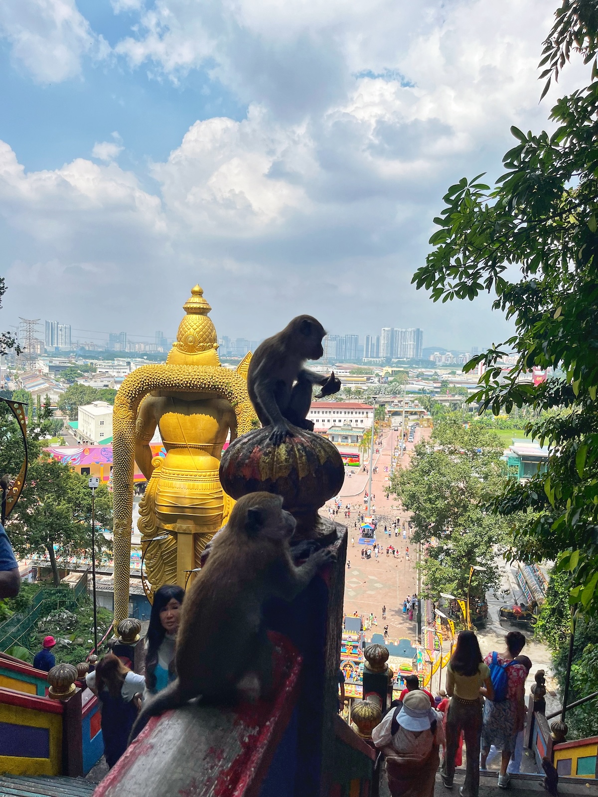 View from the Batu Caves in Malaysia, featuring two monkeys sitting on the colorful staircase railings with the golden statue of Lord Murugan and the cityscape in the background. Visitors are seen climbing the stairs, enhancing the lively atmosphere of the site.