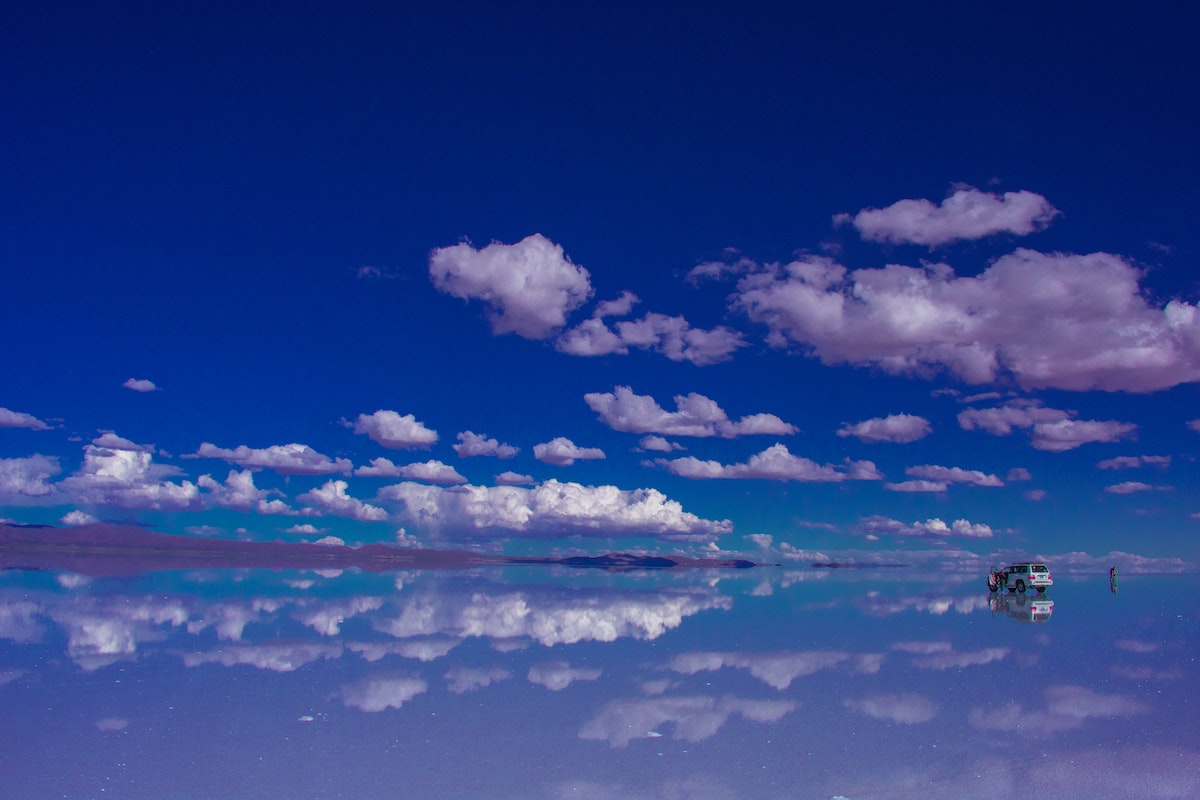 The sky reflects off the ground in a barren desert, people walk around a nearby jeep.