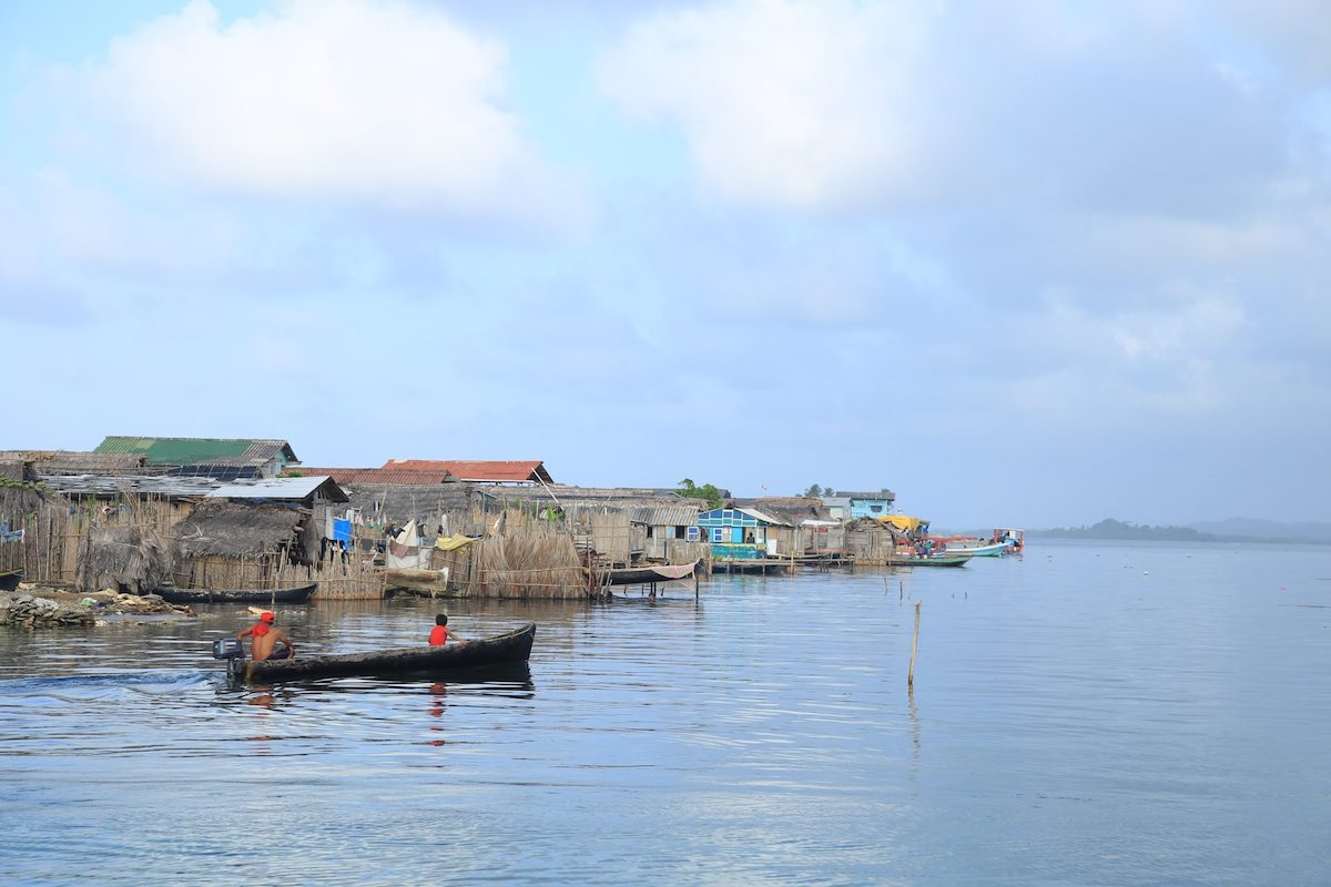 A man and a boy look onwards as they travel on a boat past wooden houses on stilts in the San Blas Islands, Panama.