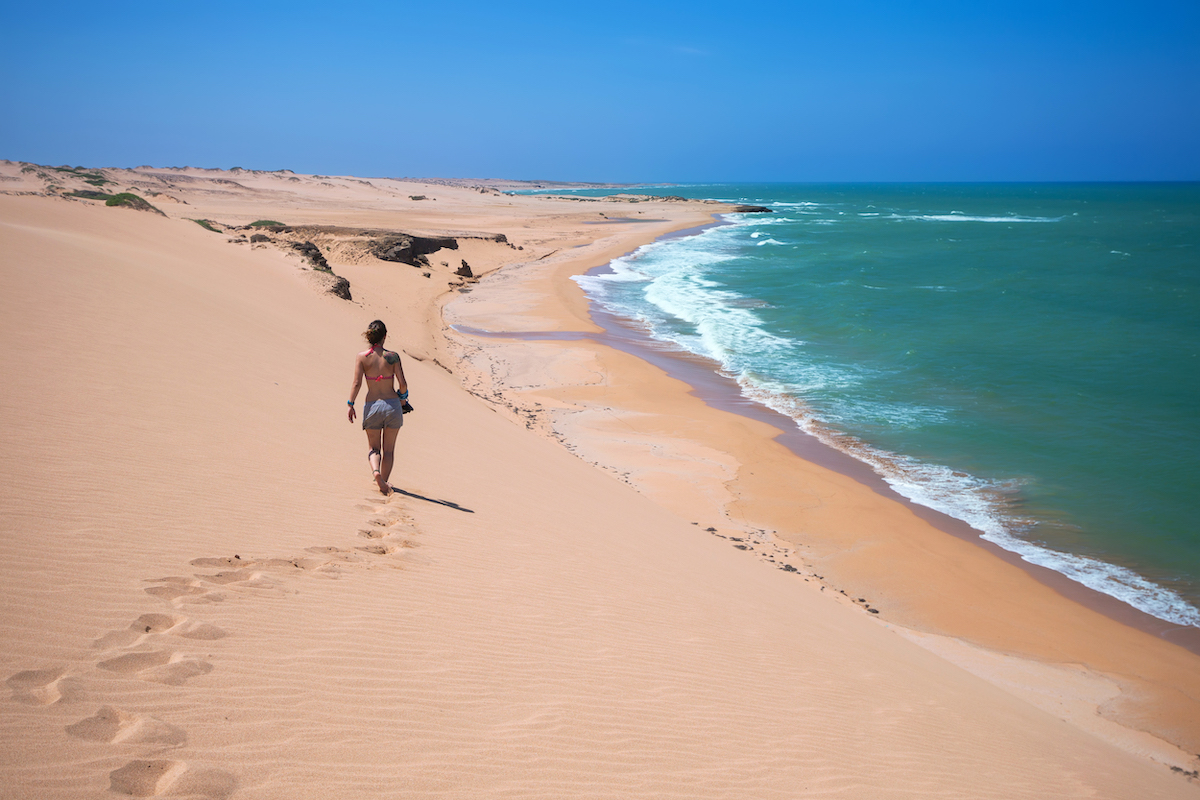 A woman walks along sand dunes alongside the shore of the torqouise sea.